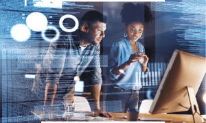 Two professionals collaborating on app development, analyzing code & digital interfaces on a computer screen in a workspace.